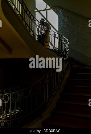 Une personne portant un chapeau se tient sur un escalier courbe, éclairé par la lumière du soleil. L'escalier a une balustrade en métal foncé avec des dessins circulaires. Banque D'Images