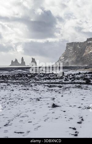 La vue des piles de basalte s'élève de façon spectaculaire de l'océan turbulent, contrastant avec la plage de sable noir enneigée sous un ciel nuageux, Vik, IC Banque D'Images