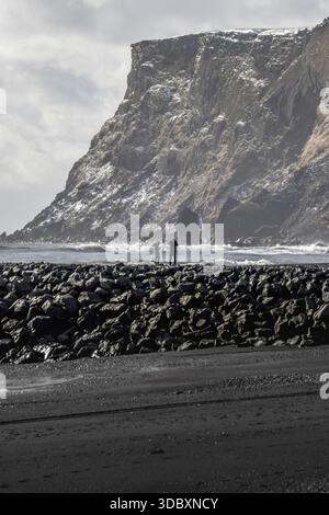 Vue de deux personnages debout sur une barrière rocheuse sombre contre la toile de fond d'une falaise massive poussiéreuse de neige, contrastant avec le sable noir BE Banque D'Images