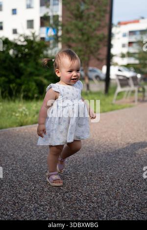 adorable tout-petit explorant joyeusement un sentier de parc, habillé dans une tenue florale mignonne avec des arbres, des bâtiments et des bancs créant un plein air urbain animé Banque D'Images