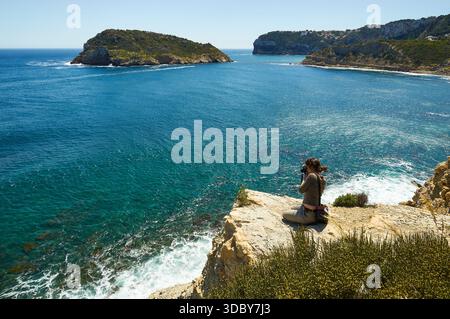 Photographe femme photographiant depuis les falaises dans le sentier de randonnée SL-CV 98, avec l'île de Portitxol en arrière-plan (Portichol, Jávea, Alicante, Espagne) Banque D'Images