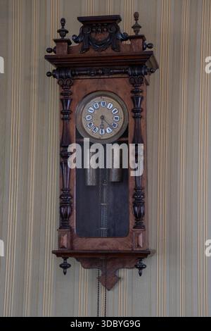 Horloge murale antique en bois avec des détails sculptés, pendule et poids montés sur papier peint rayé, tons bruns chauds et artisanat vintage Banque D'Images
