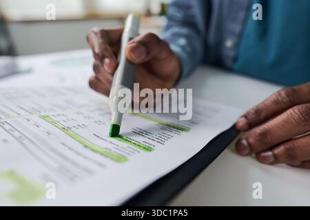 Homme noir mettant en évidence les données financières sur le document imprimé avec marqueur vert, se concentrant sur l'analyse des chiffres et la prise de notes, gros plan des mains travaillant au bureau dans le cadre du bureau Banque D'Images