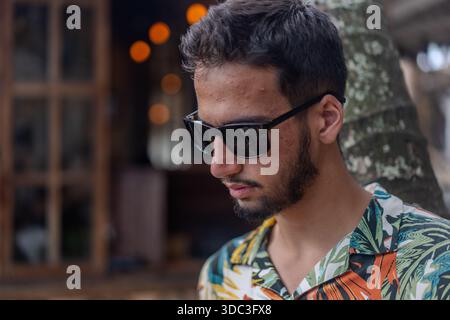 Un homme en chemise hawaïenne boit un verre avec une paille. Il est assis sous un arbre Banque D'Images