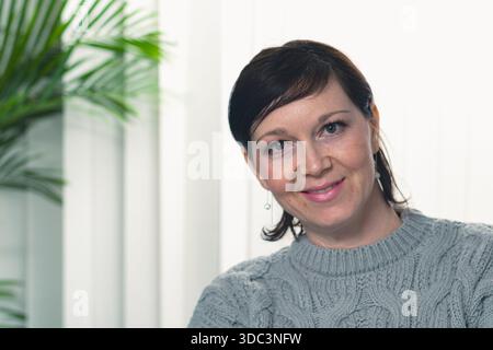 Une femme aux cheveux bruns et un pull gris sourit. Elle est assise devant une usine Banque D'Images
