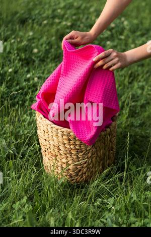 Serviettes gaufrées en coton brillant disposées dans un panier tissé sur de l'herbe verte à la lumière chaude du soleil. Cadre d'été extérieur frais, textures naturelles, confortable et propre Banque D'Images
