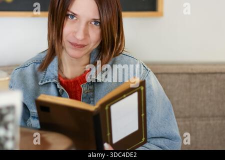 Jeune femme avec un sourire doux absorbé dans le livre tout en étant assis confortablement à l'intérieur Banque D'Images