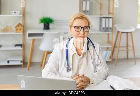 Senior souriant concentré médecin sérieuse sérieuse dans des lunettes et uniforme blanc assis au bureau en ligne Banque D'Images