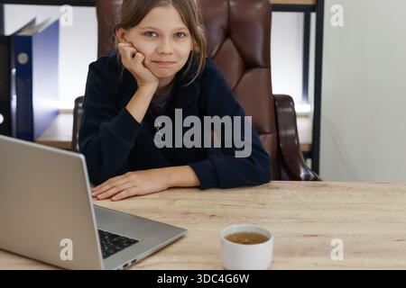 Femelle preteen ennuyée appuyée sur le coude pendant le travail à distance avec une boisson chaude à proximité Banque D'Images