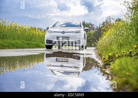 A white electric car on a country road, reflected in a puddle, surrounded by yellow flowers and blooming nature, ID3 VW, deer car sharing. Calw, Germa Banque D'Images