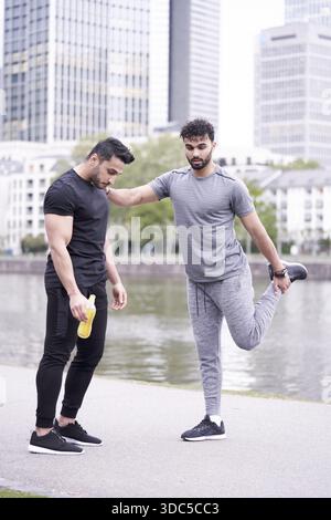 Deux copains d'entraînement s'entraînent ensemble, l'un faisant des exercices d'étirement tandis que l'autre se repose avec une bouteille en plastique d'électrolyte de boisson sportive jaune Banque D'Images
