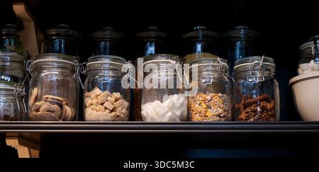 Une étagère pleine de bocaux en verre avec divers aliments à l'intérieur. Les bocaux sont disposés en rangée, certains contenant du sucre, d'autres contenant des épices, et un Banque D'Images