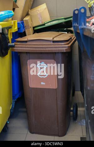 Vue avant de la poubelle à roulettes brunes avec icône de noyau de pomme et texte Bio, flanquée de conteneurs jaunes et bleus, boîtes en carton derrière Banque D'Images