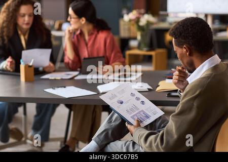 Jeune homme noir analysant les graphiques financiers tandis que deux jeunes femmes caucasiennes discutent des documents et utilisent une tablette numérique dans un cadre de bureau moderne, concept de travail d'équipe d'affaires Banque D'Images