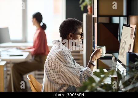 Jeune homme adulte noir portant des lunettes travaillant à l'ordinateur et tenant une tasse à café dans un bureau moderne, jeune femme adulte caucasienne travaillant au bureau en arrière-plan, les deux se sont concentrés sur les tâches Banque D'Images