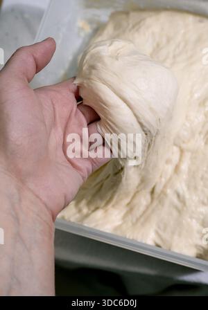 Fermez la main de la femme pétrissant la pâte à pain crue dans un récipient en plastique. Procédé de cuisson maison. Préparation élastique de pâte levante pour la cuisson au four. Banque D'Images