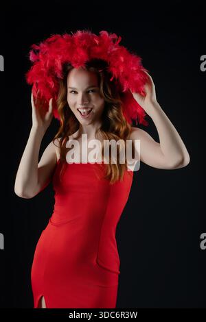 Une femme se tient dans un studio, souriant et posant avec une énergie ludique. Elle porte une robe rouge frappante et un grand chapeau à plumes, exprimant la joie et la confiance dans son expression et sa position. || autorisation du modèle Banque D'Images