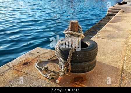 Gros plan d'une borne d'amarrage rouillée enveloppée d'une corde nautique épaisse et de vieux pneus sur une jetée de pierre ensoleillée contre l'eau de mer bleue. Banque D'Images