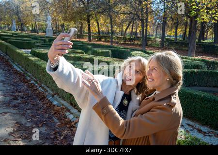Deux femmes partagent un moment joyeux en prenant des selfies dans un jardin pittoresque rempli de feuilles d'automne vibrantes. Le soleil brille brillamment, mettant en valeur leur ha Banque D'Images