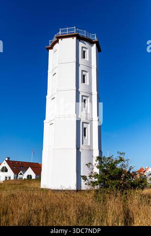 Skagen, Danemark-8 août 2025 : phare blanc lumineux avec des maisons à toit rouge en arrière-plan, debout haut sur un paysage herbeux sec sous un s bleu vif Banque D'Images