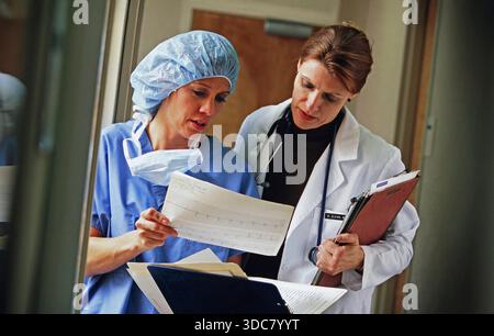 Deux professionnels de la santé discutent des dossiers de patients dans un couloir hospitalier. L'un porte des gommages et une casquette chirurgicale, tandis que l'autre est en coa blanc Banque D'Images