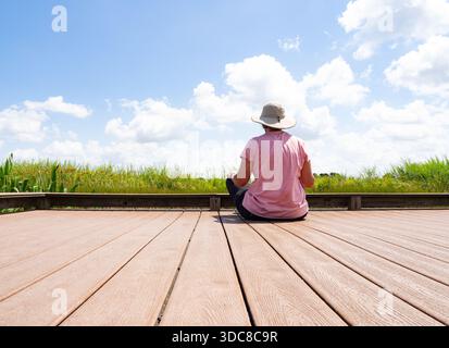 Femme âgée portant un t-shirt rose assis sur un quai surplombant un étang dans la réserve Indiangrass de la Coastal Prairie Conservancy à Waller, Texas Banque D'Images