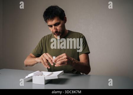 Client masculin déballant les nouveaux écouteurs sans fil achetés, retirant de la boîte sur la table grise, exprimant son enthousiasme et sa satisfaction Banque D'Images