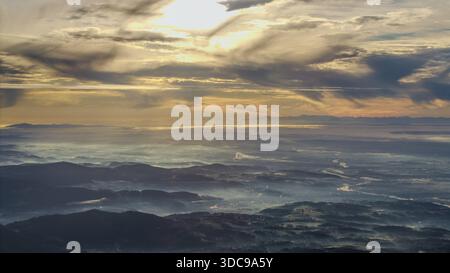 Le soleil se lève sur les montagnes. Le brouillard se répand à travers le paysage. Des nuages pendent dans le ciel. La journée commence dans la nature. Au premier plan, la ville Banque D'Images