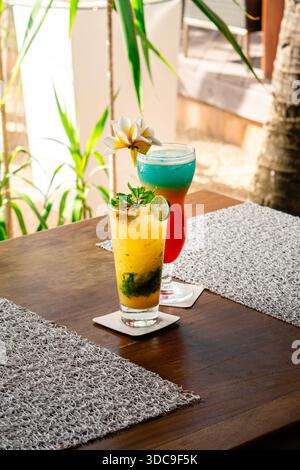 Deux cocktails tropicaux rafraîchissants, de couleur ambre et turquoise, sont assis sur une table rustique en bois. Garni de citron vert, de menthe et d'une fleur délicate. Banque D'Images