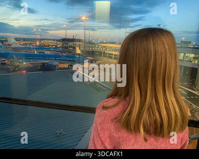 Un enfant regarde par une fenêtre du terminal de l'aéroport au-dessus de l'avion à l'aéroport d'Amsterdam Schiphol au crépuscule. Amsterdam, pays-Bas, novembre 2024 Banque D'Images