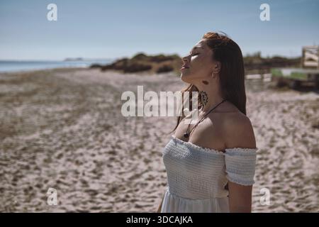 Jeune femme sereine en robe blanche profiter de la chaleur et du temps ensoleillé avec les yeux fermés se tient sur la plage de sable, se sentir libre et heureux. Moment méditatif Banque D'Images