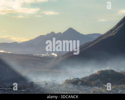 Vue aérienne du brouillard bas tissant à travers les vallées et au-dessus de la ville sous la majestueuse chaîne de montagnes sombre sous un ciel pastel, San Michele di Seri Banque D'Images