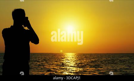 Une photo de silhouette de haute qualité montrant l'homme prenant des photos de la mer au coucher du soleil pendant un coucher de soleil dramatique. La scène capture l'émotion, le style de vie, le freedo Banque D'Images