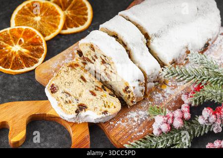 Traditionnel stollé de Noël, pain de fruits sucrés tranché sur une planche de bois, saupoudré de sucre en poudre sur fond texturé noir. Épices, fruits secs Banque D'Images