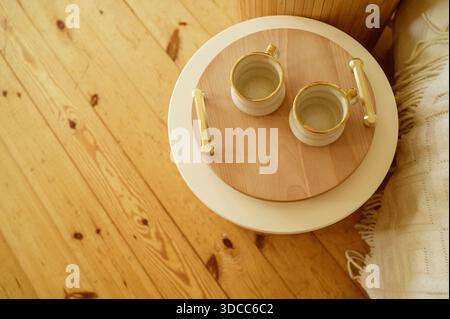 Deux mugs en céramique blanche avec bords dorés sur un plateau rond en bois avec poignées dorées. Concept de matin confortable, style de vie hygge et maison de vacances minimaliste Banque D'Images