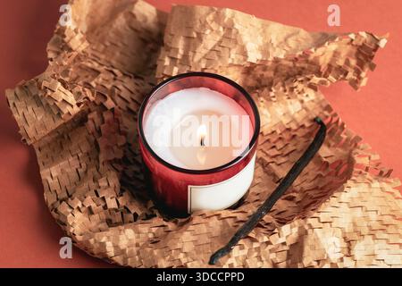 Bougie parfumée allumée dans un pot en verre avec gousse de vanille enveloppée dans du papier kraft texturé sur fond orange chaud. Vue de dessus. Banque D'Images