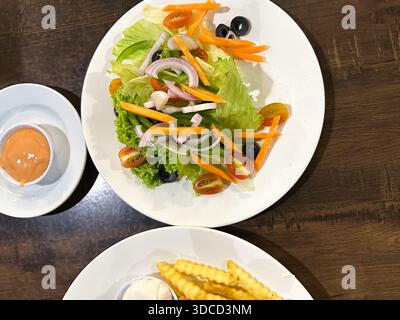 Une salade de légumes colorée avec laitue, tomates cerises, carottes, oignons rouges et olives noires servies sur une assiette blanche dans un cadre chaleureux. Banque D'Images