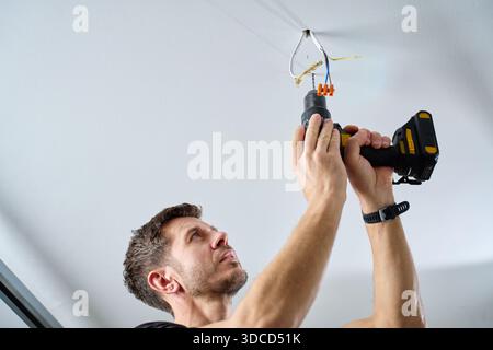 L'homme portant une combinaison de travail perce un trou dans le plafond à l'aide d'une perceuse électrique sans fil dans la pièce intérieure. Concept de rénovation domiciliaire, d'installation et professionnelle Banque D'Images