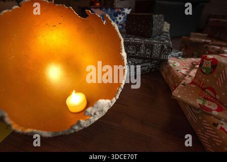 Gros plan d'une bougie incandescente à l'intérieur d'un bol décoratif avec des cadeaux de Noël emballés en arrière-plan, créant une ambiance de vacances chaleureuse et intime. Banque D'Images