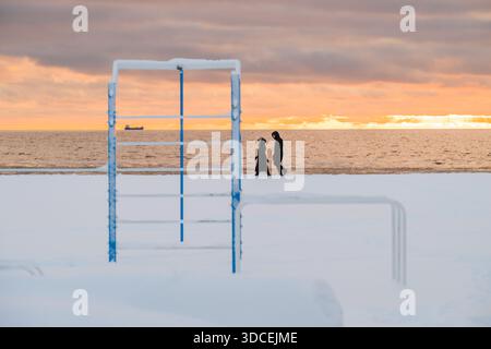 Vue d'un couple de promenades le long de la plage enneigée alors que le soleil se couche sur la mer Baltique, projetant une lueur chaude sur l'horizon, Parnu, Estonie. Banque D'Images