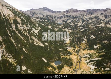 Vue aérienne des sommets escarpés et des pentes verdoyantes rencontrant des lacs sombres et encore sous un ciel sombre dans les montagnes Pirin, Bansko, Bulgarie. Banque D'Images