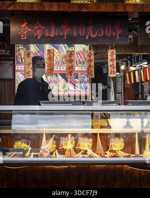 Fenghuang, Chine - 24 mars 2024 : vue d'un vendeur en tenue traditionnelle se tient derrière une exposition de nourriture de rue vibrante, baignée dans la lueur chaude de ov Banque D'Images