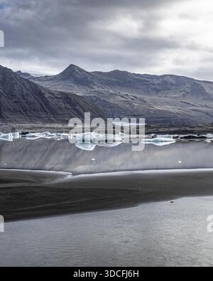 Vue sur les sables noirs et les eaux glacées reflétant le ciel discret et les imposantes montagnes au loin, Islande. Banque D'Images