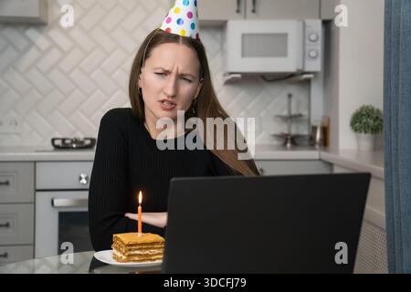 Femme triste portant chapeau de fête célébrant l'anniversaire en ligne pendant la pandémie, faisant un appel vidéo avec un ordinateur portable et regardant un petit gâteau avec une bougie allumée Banque D'Images