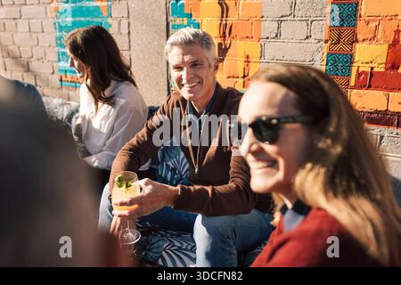 Homme heureux tenant un verre et parlant avec des amis tout en étant assis à l'extérieur du bar à la journée ensoleillée Banque D'Images