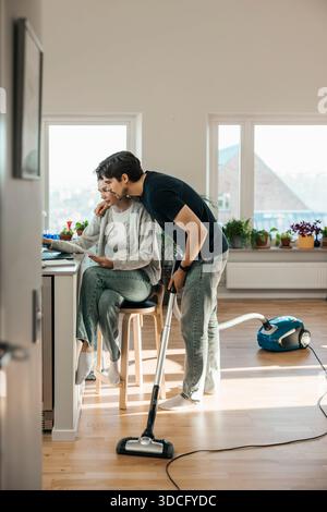Jeune homme tenant aspirateur avec bras autour de la petite amie assise à table pour la tenue de livres à la maison Banque D'Images