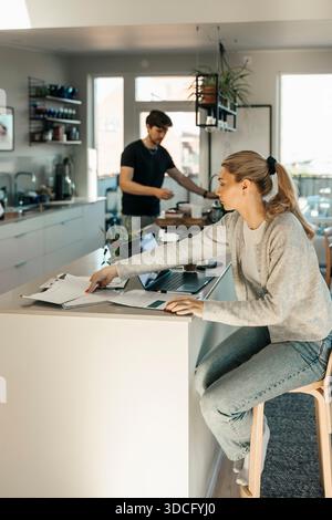 Jeune femme examinant des documents financiers tout en étant assise à l'îlot de cuisine à la maison Banque D'Images