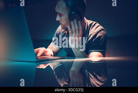 Un homme utilise un ordinateur portable tout en portant des écouteurs dans une pièce sombre pendant la nuit. Banque D'Images