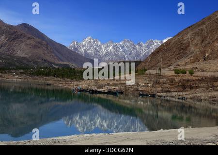 Vue sur le lac serein reflétant les montagnes enneigées sous un ciel bleu vif, encadré par des collines brunes accidentées à Passu, Gilgit Baltistan, Pakistan Banque D'Images