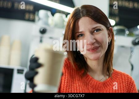 Jeune femme ludique avec des cheveux auburn dans un pull vibrant étend un café à emporter dans un environnement de café animé. Banque D'Images